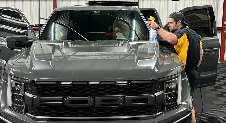 Black Optix Tint technician installing window tint on a gray Ford F-150 Raptor inside the shop, prepping glass for a smooth, bubble-free finish.