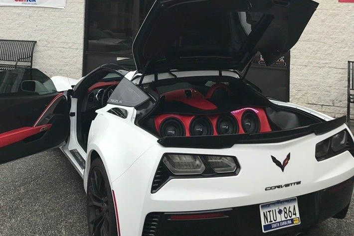 White Chevrolet Corvette with doors and rear hatch open, showcasing a custom red-and-black trunk audio system installation at Black Optix Tint.